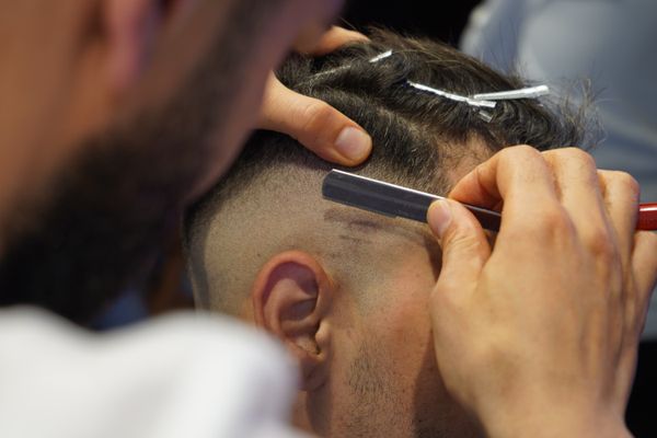 Close view over the barber's shoulder: the top sectioned with clips while the razor squares the line behind the ear.