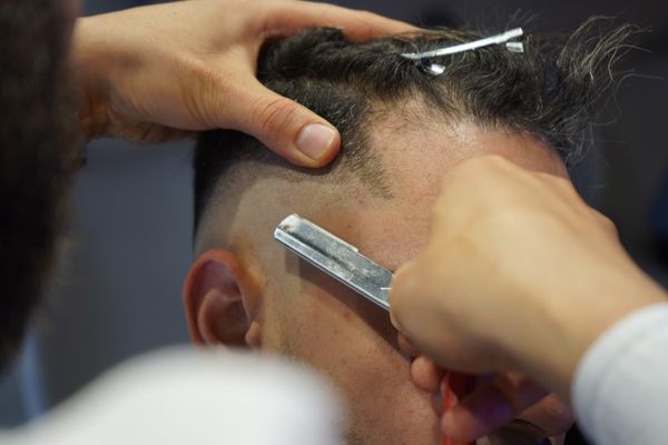 A barber steadies a client's head with one hand while working a straight razor along the faded edge above the ear.