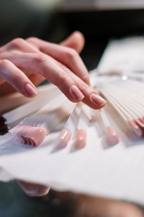 A hand with soft nude gel nails resting beside a fan of colour swatch sticks in natural window light, close enough that the cuticle line and the sidewalls are visible.